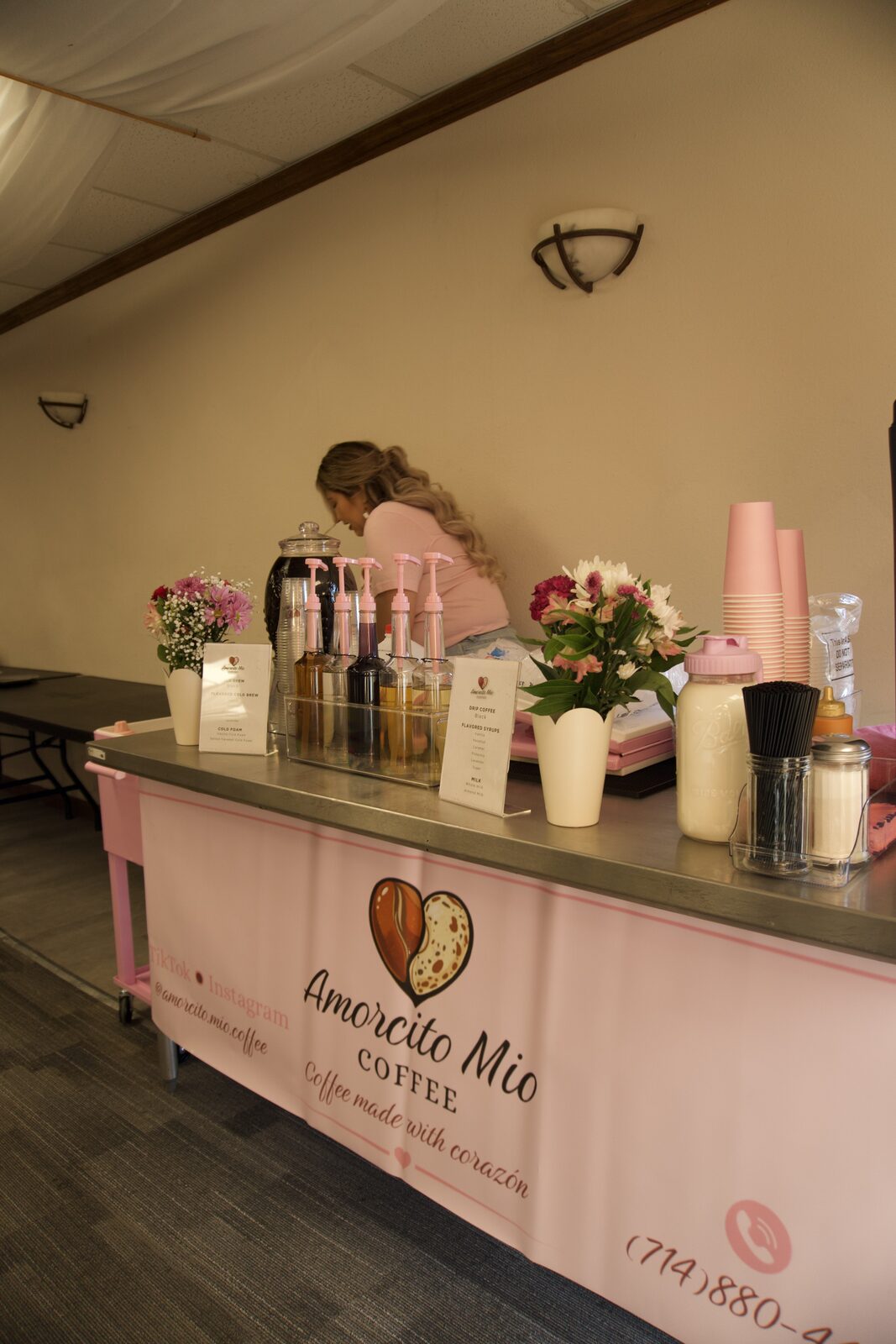 The pink Amorcito Mío coffee cart set up indoors with fresh flowers, syrup bottles and pink cups.