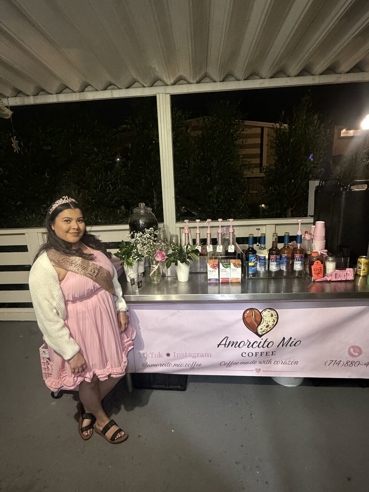 A quinceañera in a pink dress standing beside the Amorcito Mío cart at her party.